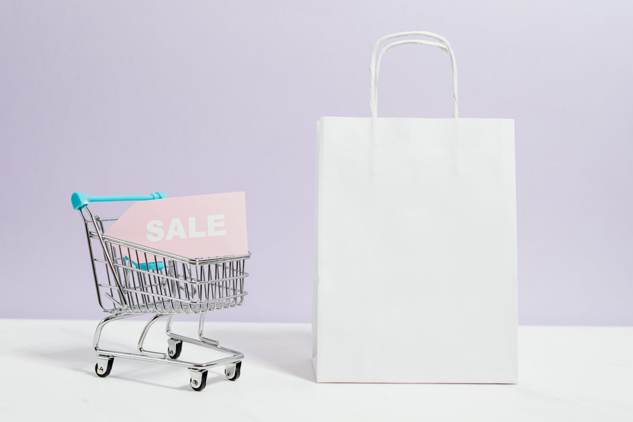 A miniature shopping cart with a sale sign beside a paper bag, perfect for online shopping themes.